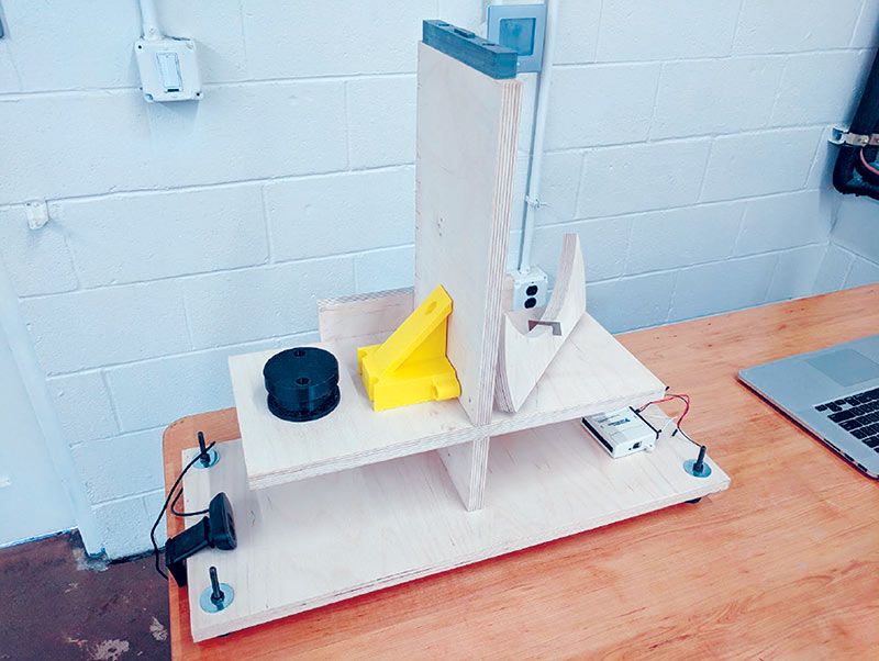 The prototype components ­‑ wood frame, pivot mount (gray), coil housing (black), support piece for the nylon guides (yellow), balance wheel and razor-blade pivot (resting upside down on the right side of the wooden frame), webcam (far left), and data acquisition device (far right). Photo courtesy of John Evans.